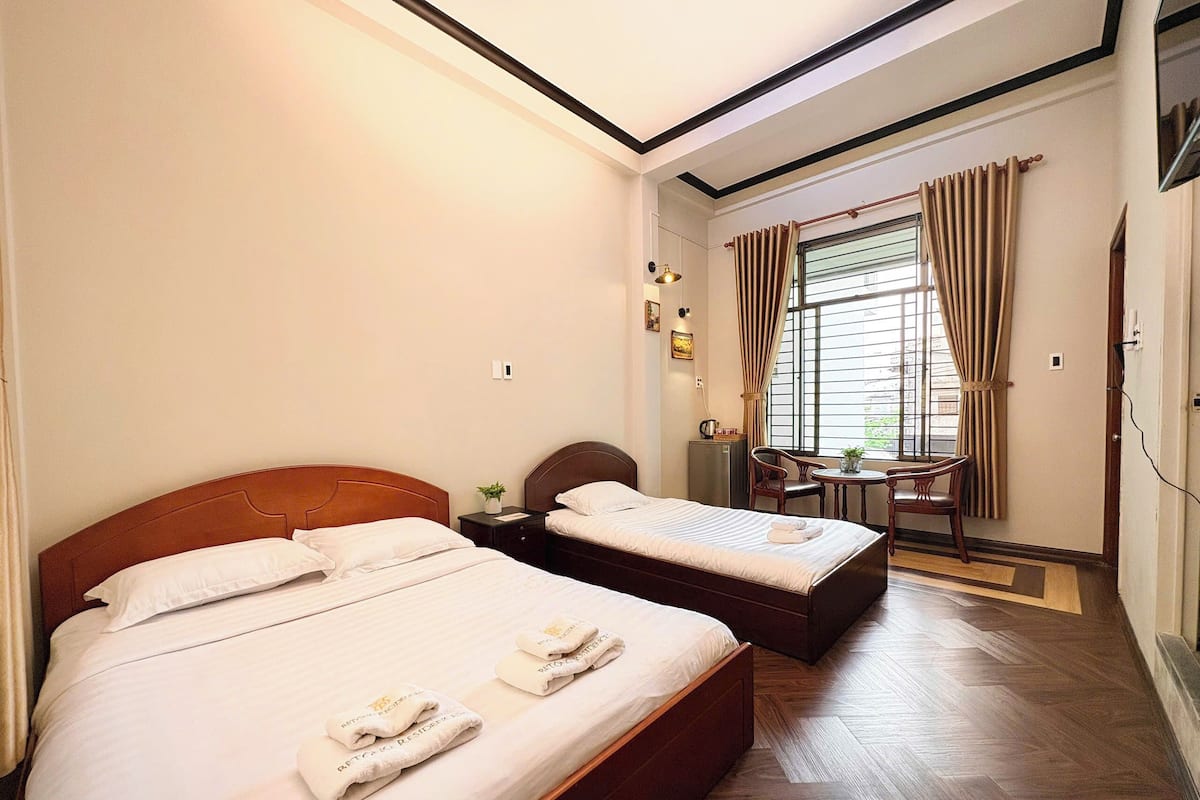 A hotel room with two beds, a small table, a chair, and a window with curtains.