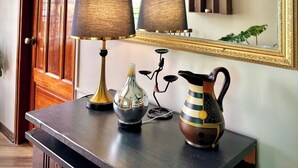 A hallway with a wooden door, a dark wooden console table with a lamp, a vase, and a pitcher. A large mirror reflects a chandelier and a window. - Bê Tông Residences Homestay (Can Tho)