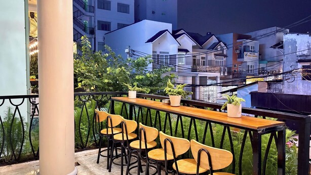 An outdoor seating area with a wooden table and chairs, surrounded by greenery and overlooking a residential area with buildings. - Bê Tông Residences Homestay (Can Tho)