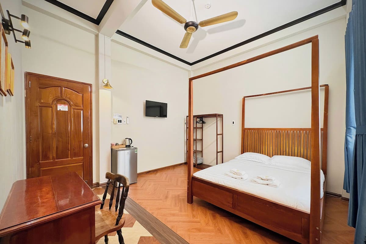 A bedroom with a four-poster bed, a wooden desk, a chair, a television, a wardrobe, and a ceiling fan.