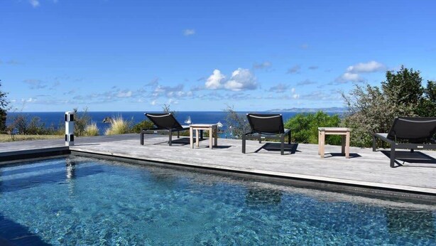 A clear pool with lounge chairs and a table, overlooking a scenic view of the ocean.
