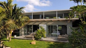 Una casa moderna de dos pisos con techo plano, amplios ventanales y un jardín cuidado con palmeras.
