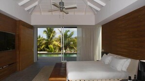 Un dormitorio con una cama grande, ventilador de techo, televisor y vista a palmeras a través de una puerta corrediza de vidrio.