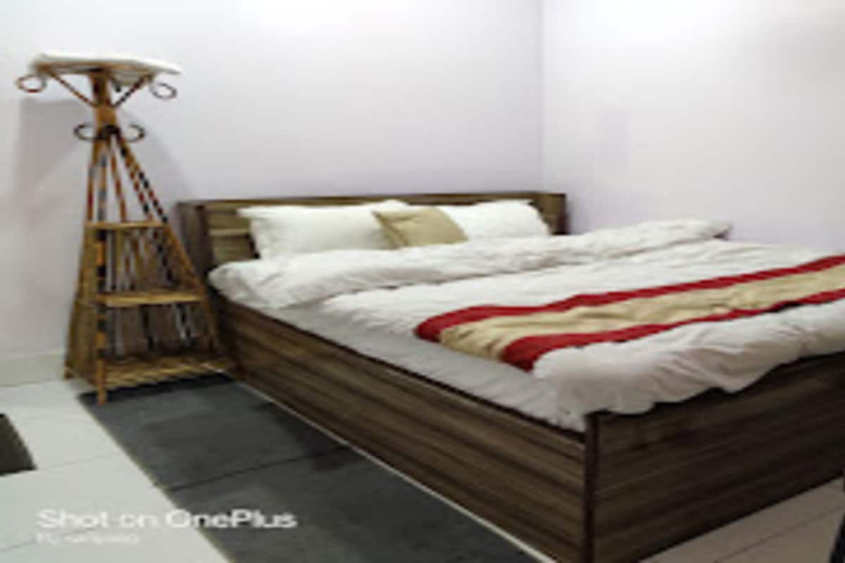 Una cama individual con estructura de madera, ropa blanca de cama y una colcha a rayas rojas y blancas. Hay una mesita de noche y una lámpara de pie.