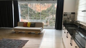 Een moderne woonkamer met een bank, een groot raam met uitzicht op een bergachtig landschap, een keuken met zwarte aanrechtbladen en een vloerkleed met een patroon.