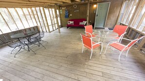 Eine offene Holzterrasse mit einem runden Glastisch und orangefarbenen Stühlen, einer roten Couch und einer Tür im Hintergrund.