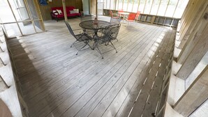 Eine überdachte Holzterrasse mit einem runden Metalltisch und Stühlen, einem roten Sofa und einer Tür, die nach draußen führt.