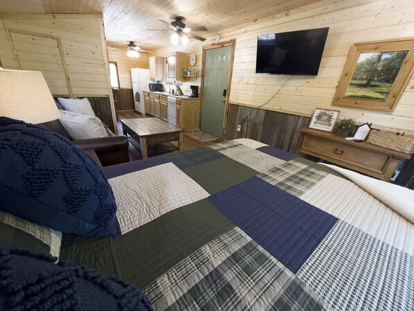 A cozy cabin interior with a bed, a kitchenette, a dining area, and a TV mounted on the wall.
