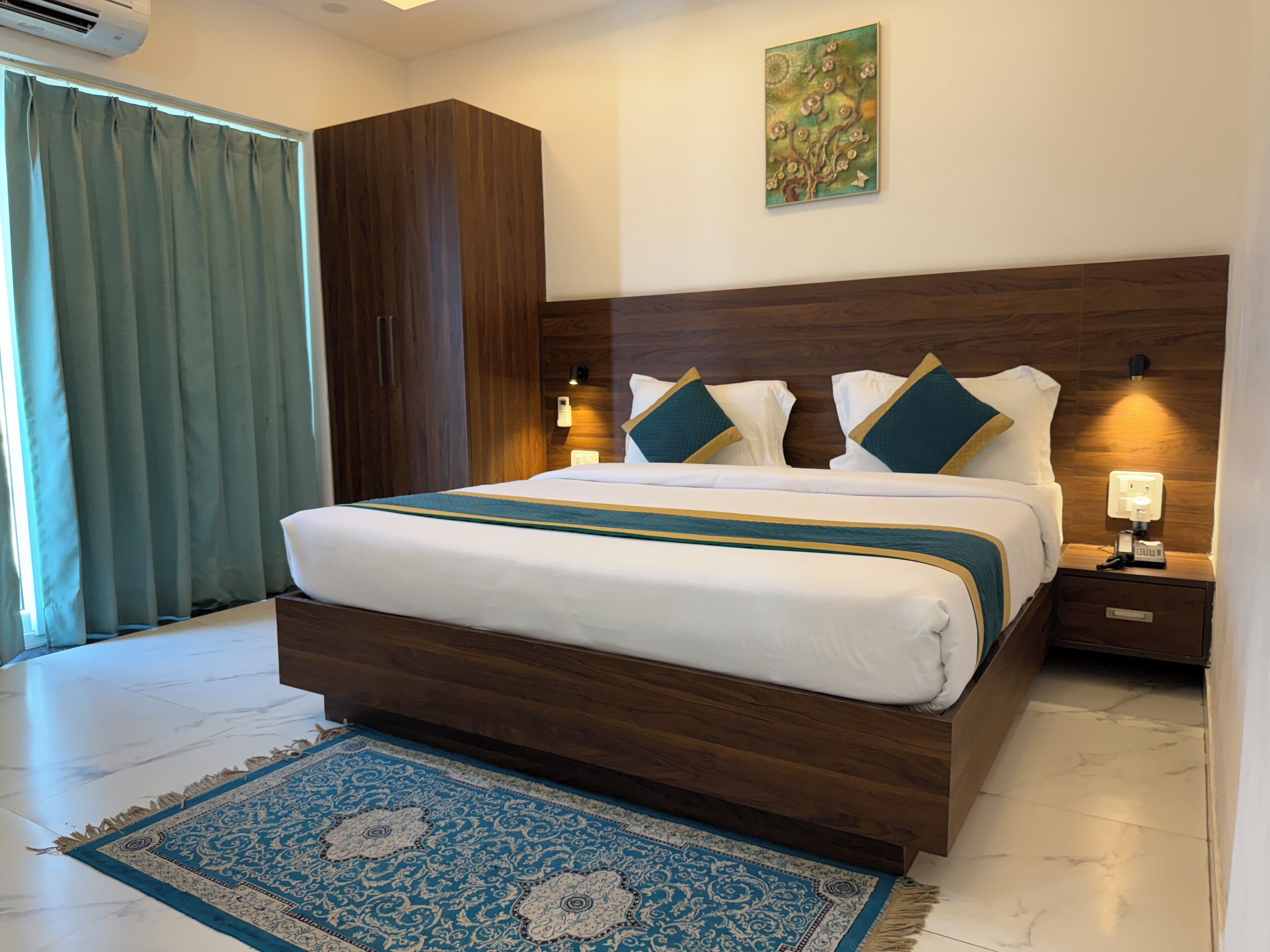 A hotel room with a large bed, wooden headboard, two bedside lamps, a nightstand with a phone, and a blue patterned rug.