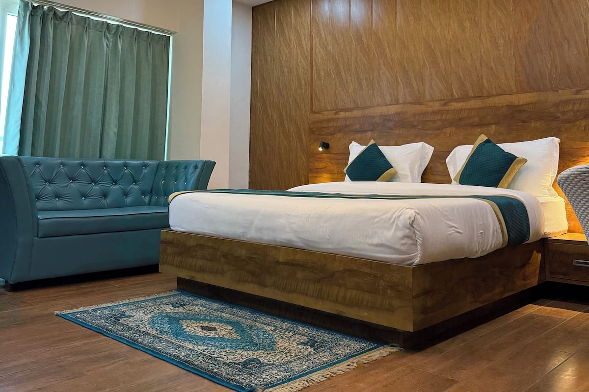 A modern hotel room with a wooden headboard, a tufted blue sofa, a large bed with white bedding and teal pillows, a blue patterned rug, and a ceiling fan.