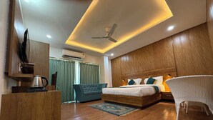 Une chambre d’hôtel moderne avec un plancher en bois, un grand lit avec une literie verte et blanche, un canapé, un fauteuil, un bureau avec une bouilloire et un ventilateur de plafond.