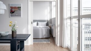 Un dormitorio moderno con un ventanal grande, una cama con ropa de cama blanca, una mesita de noche con un cuenco y flores, y un cuadro en la pared.