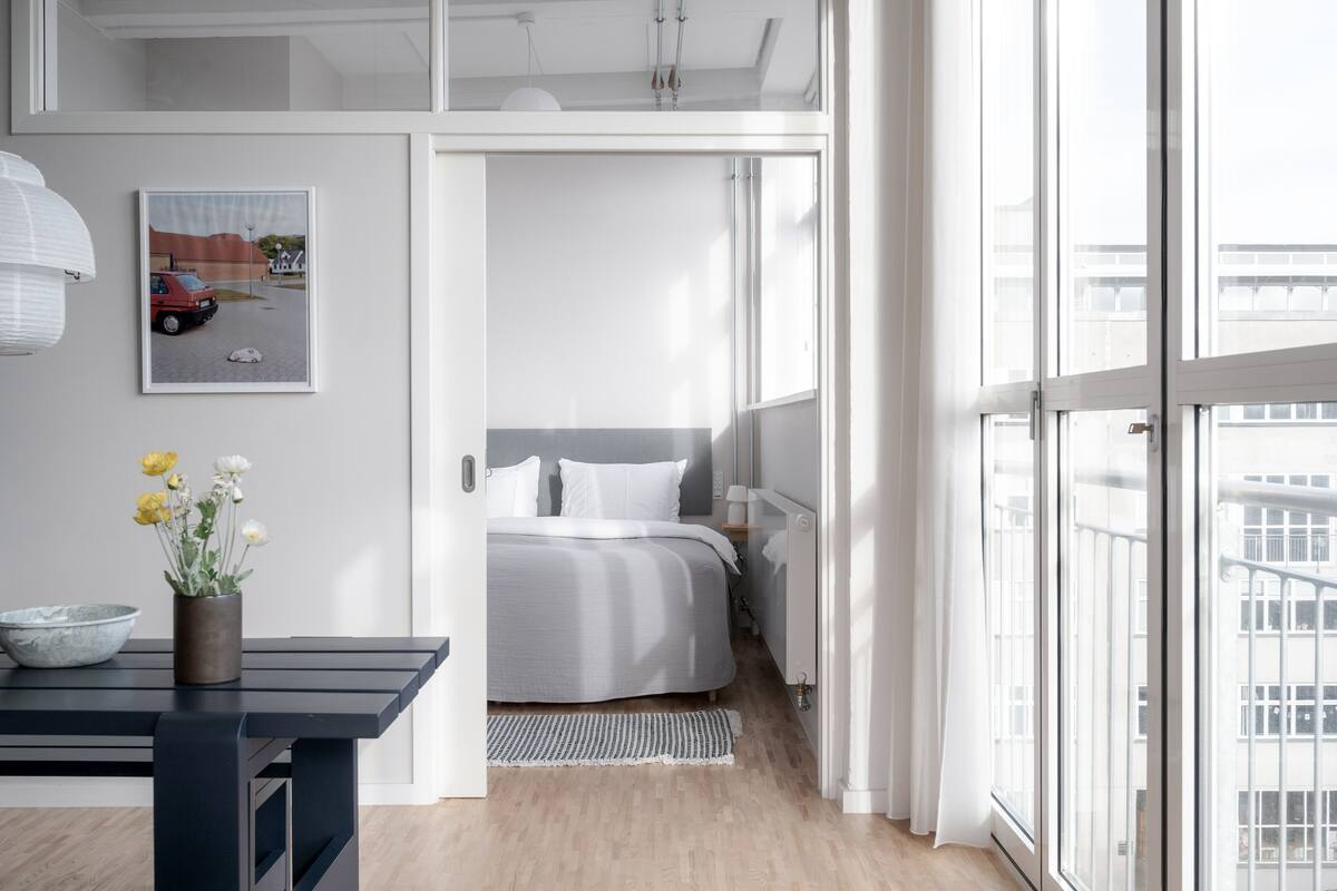A modern bedroom with a large window, a bed with white bedding, a bedside table with a bowl and flowers, and a framed picture on the wall.