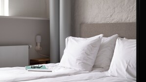 Una cama bien hecha con ropa blanca y almohadas, una mesita de noche con un libro y gafas, y un teléfono fijado a la pared.