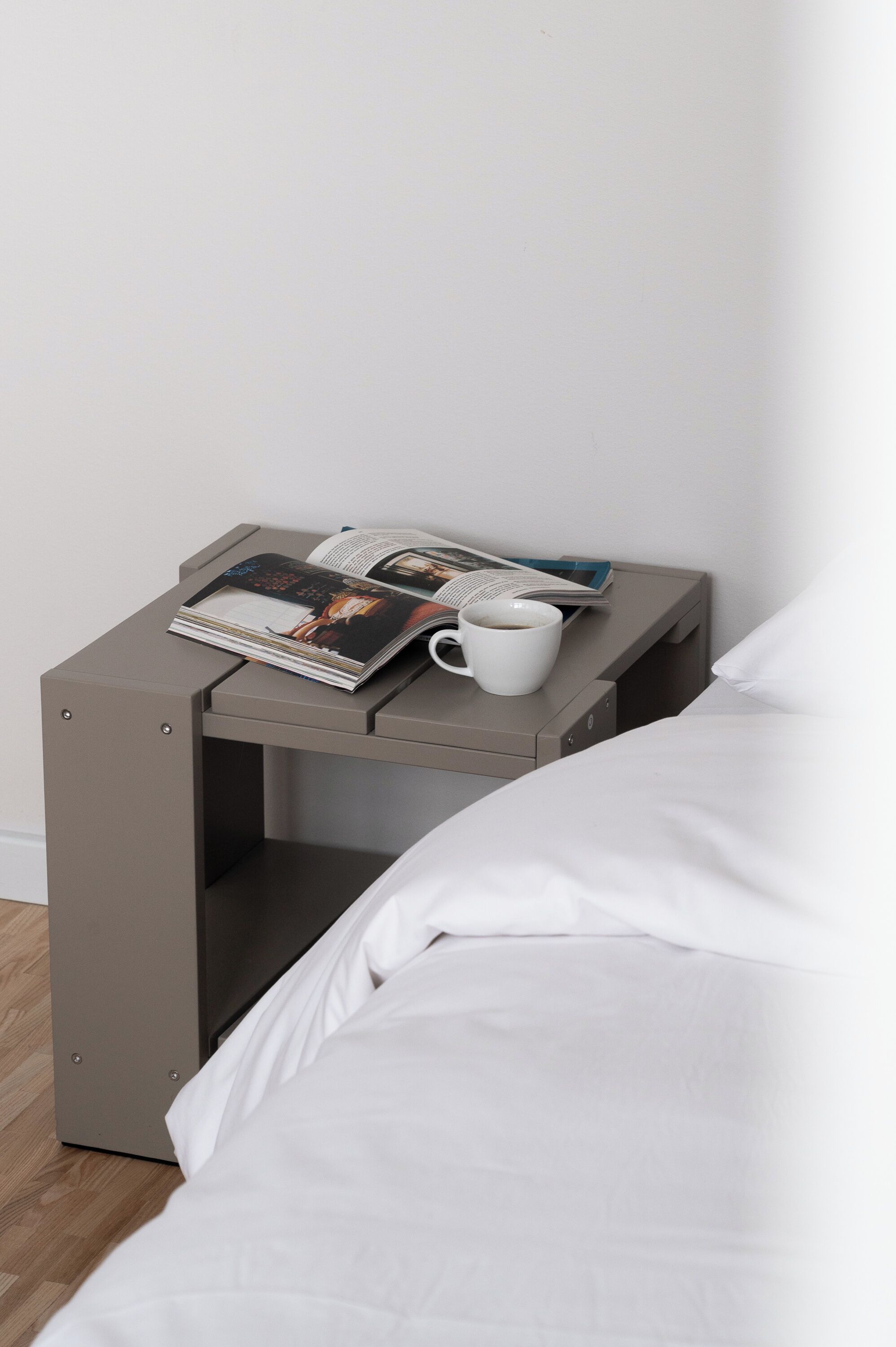 A neatly made bed with white sheets and a small bedside table with a magazine, a book, and a white mug.