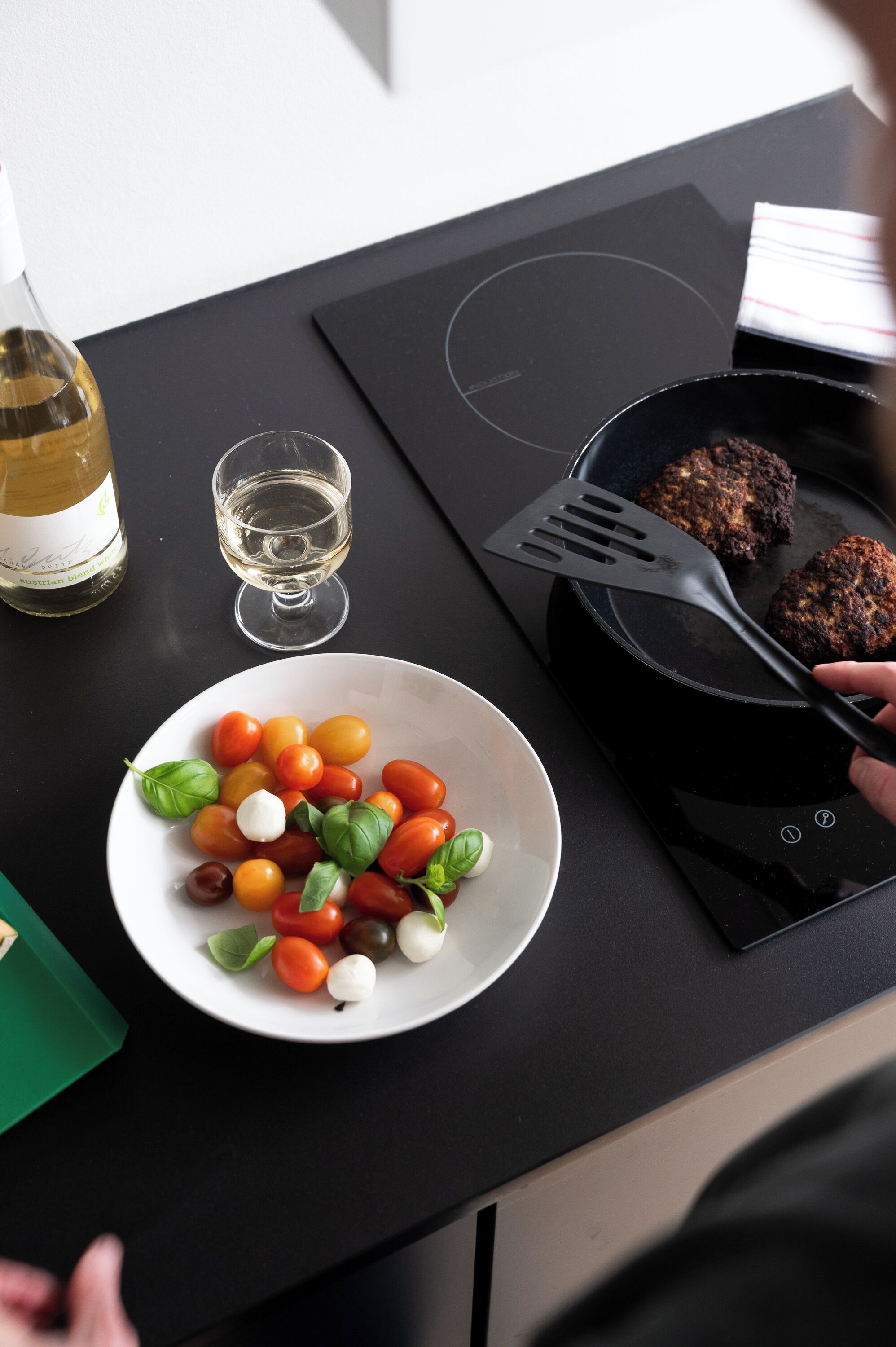 A kitchen counter with a plate of cherry tomatoes, a glass of white wine, and a frying pan with meat patties.