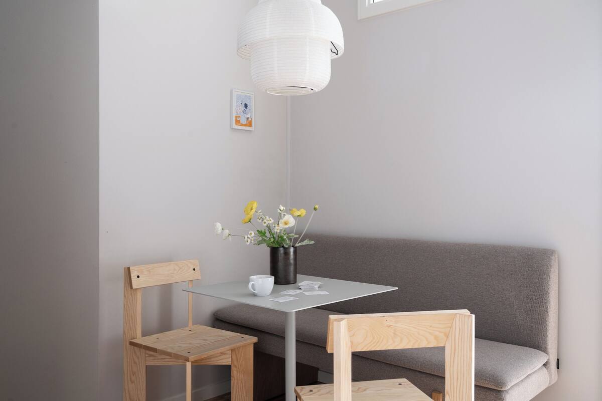 A minimalist room with a grey sofa, two wooden chairs, a small table, a white pendant light, and a vase with flowers.