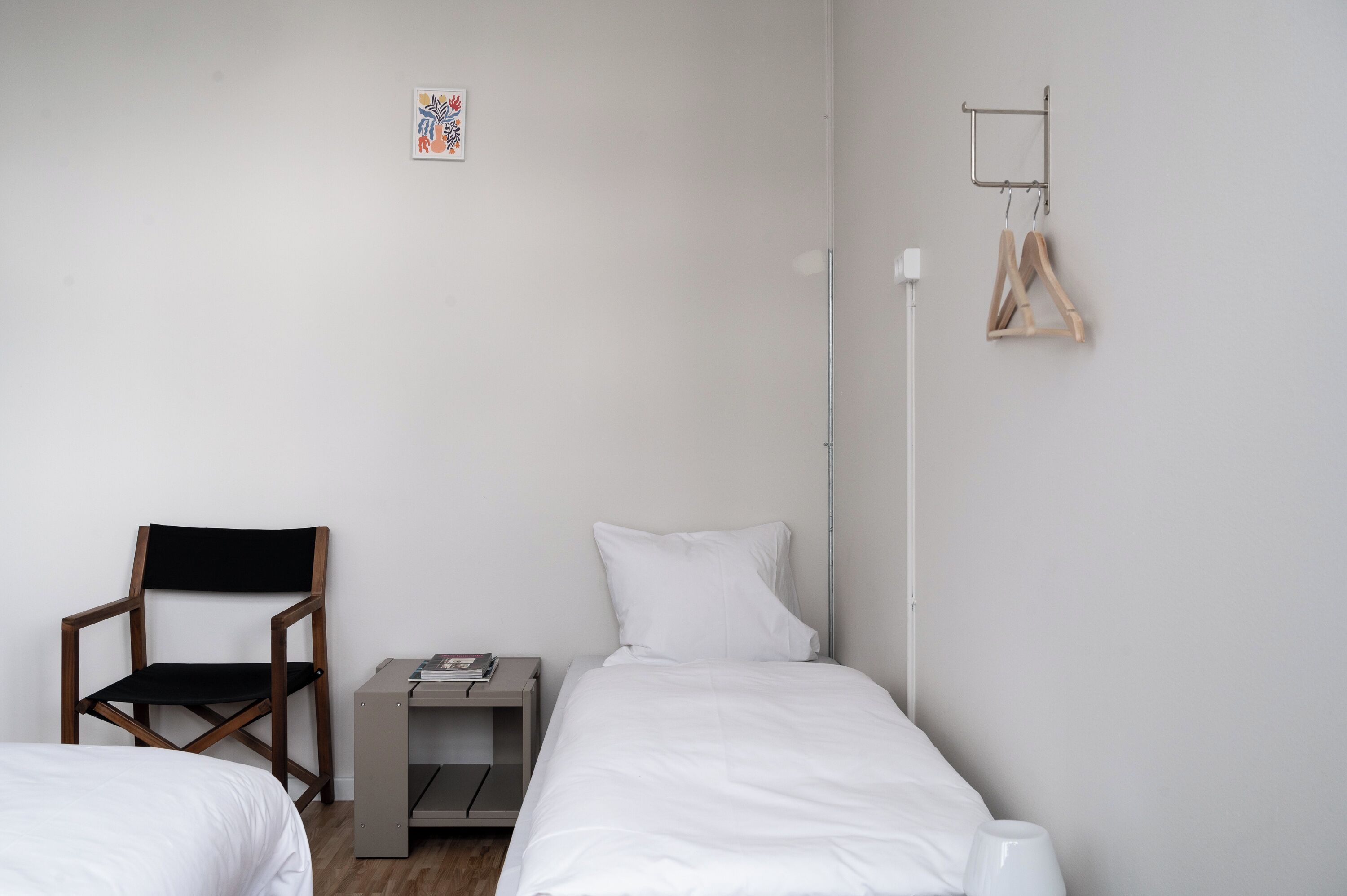 Un dormitorio con dos camas individuales, una silla de madera, una mesita de noche con un libro y un toallero en la pared.