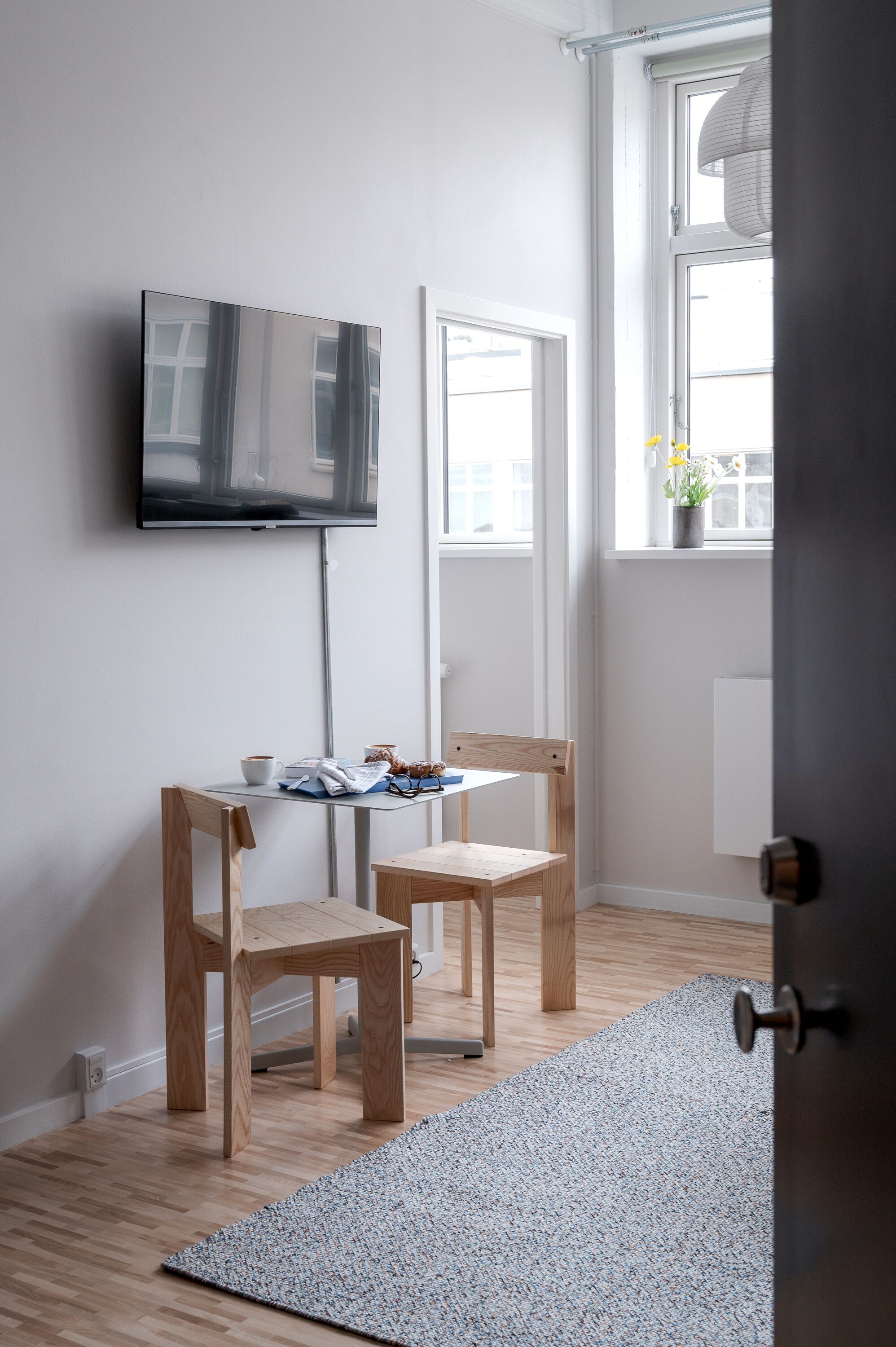 Una habitación con televisión montada en la pared, dos sillas de madera, una mesita con una taza y un libro, una ventana con vistas al exterior y una puerta.