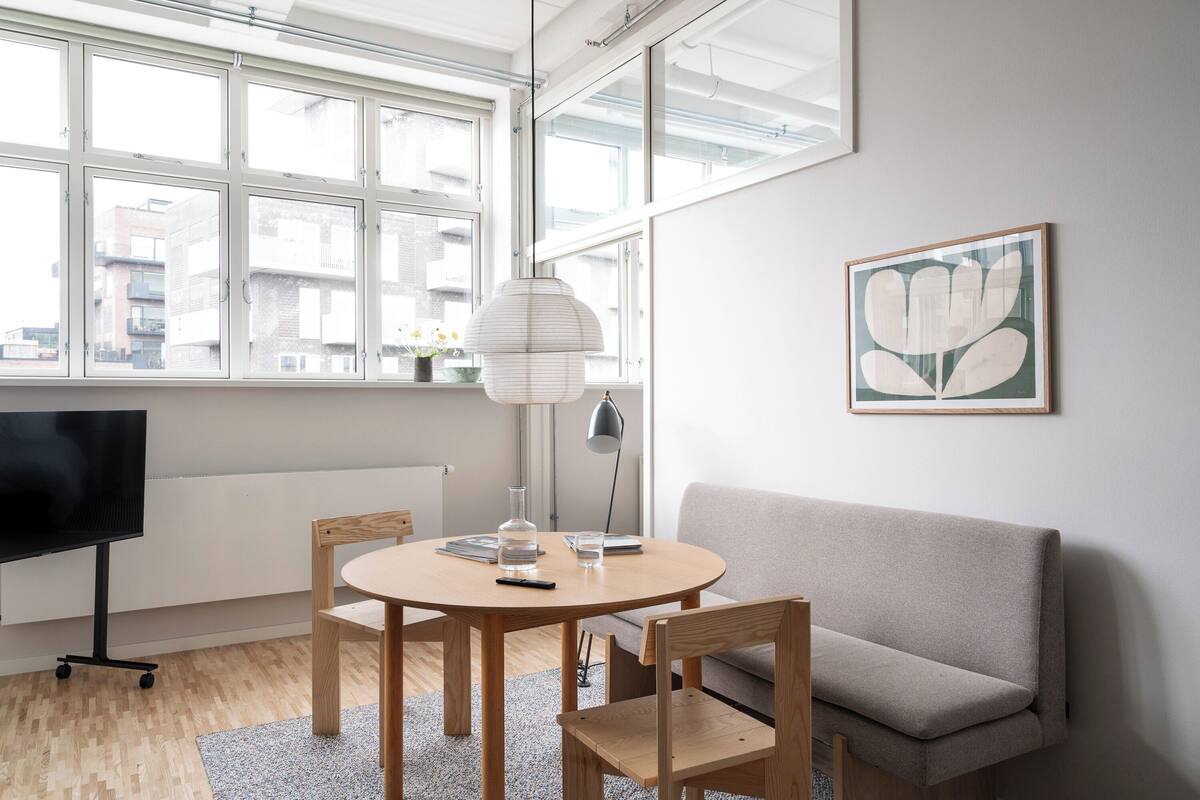 A modern living room with a round wooden table, two chairs, a grey sofa, a TV on a stand, a lamp, and a framed artwork on the wall.