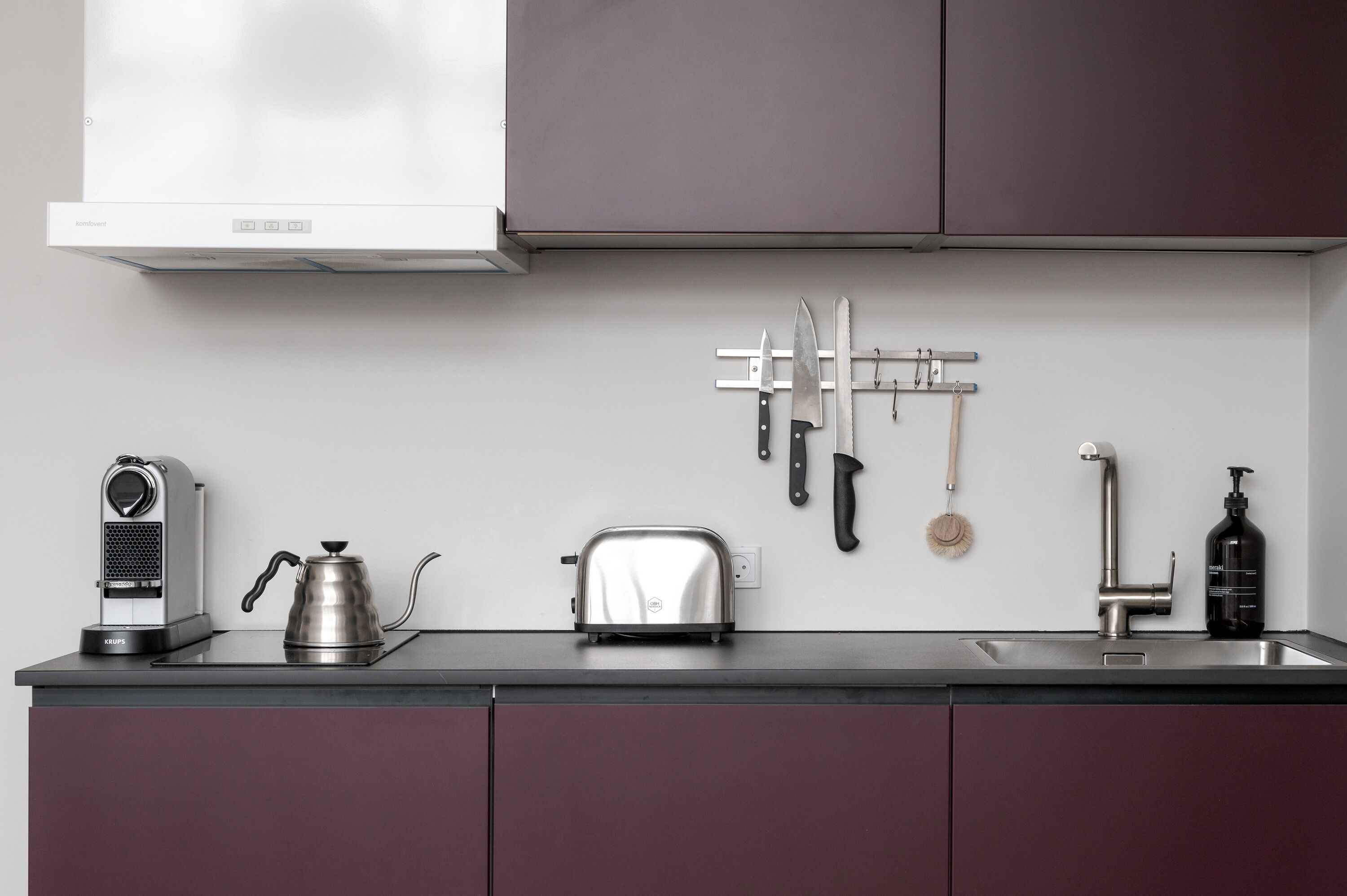 A kitchen with a toaster, kettle, and coffee maker on the counter, a knife rack on the wall, and a sink with a soap dispenser.