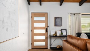 Ein Zimmer mit Holztür, einem schwarzen Konsolentisch mit gerahmten Gegenständen, einer braunen Ledersofa und einem Fenster mit Jalousien.