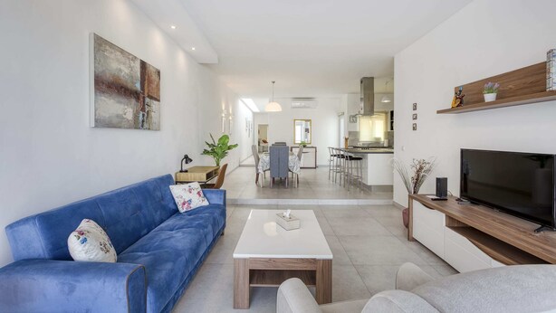 A modern living room with a blue sofa, a white coffee table, and a grey sofa. There is a TV on a wooden stand and a kitchen in the background. - Gzira Penthouse (Gzira)