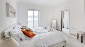 A modern bedroom with a large bed, a bedside table, a chair, and a window with a city view. - Gzira Penthouse (Gzira)