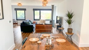 A spacious living area with a wooden dining table set for six, a sofa, a fireplace, and large windows offering a view of the outdoors.