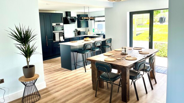 A modern kitchen with dark cabinetry, a central island, and a dining area with wooden tables and chairs.