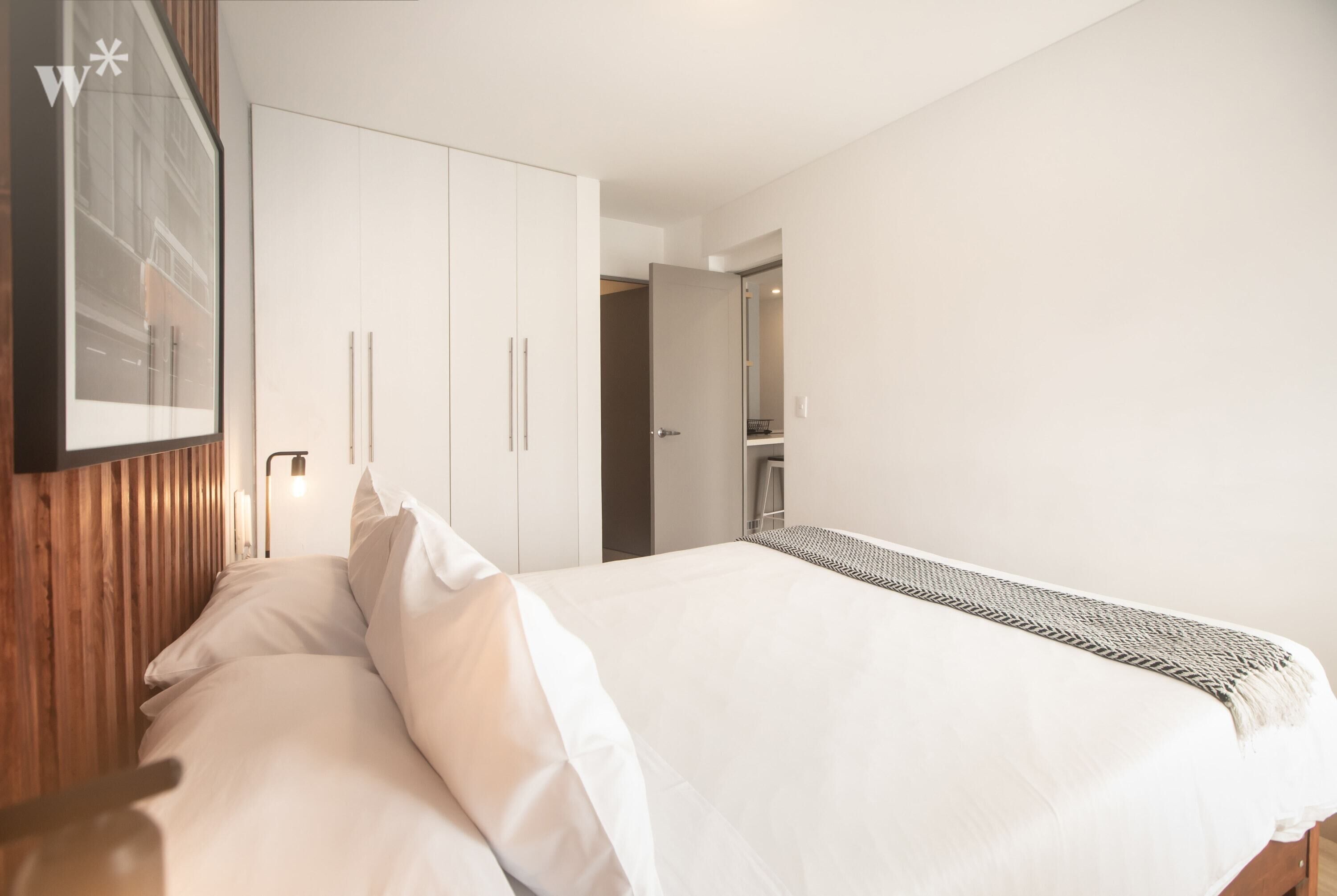 Un dormitorio moderno con una cama grande, cabecera de madera, armario blanco y un cuadro en la pared.