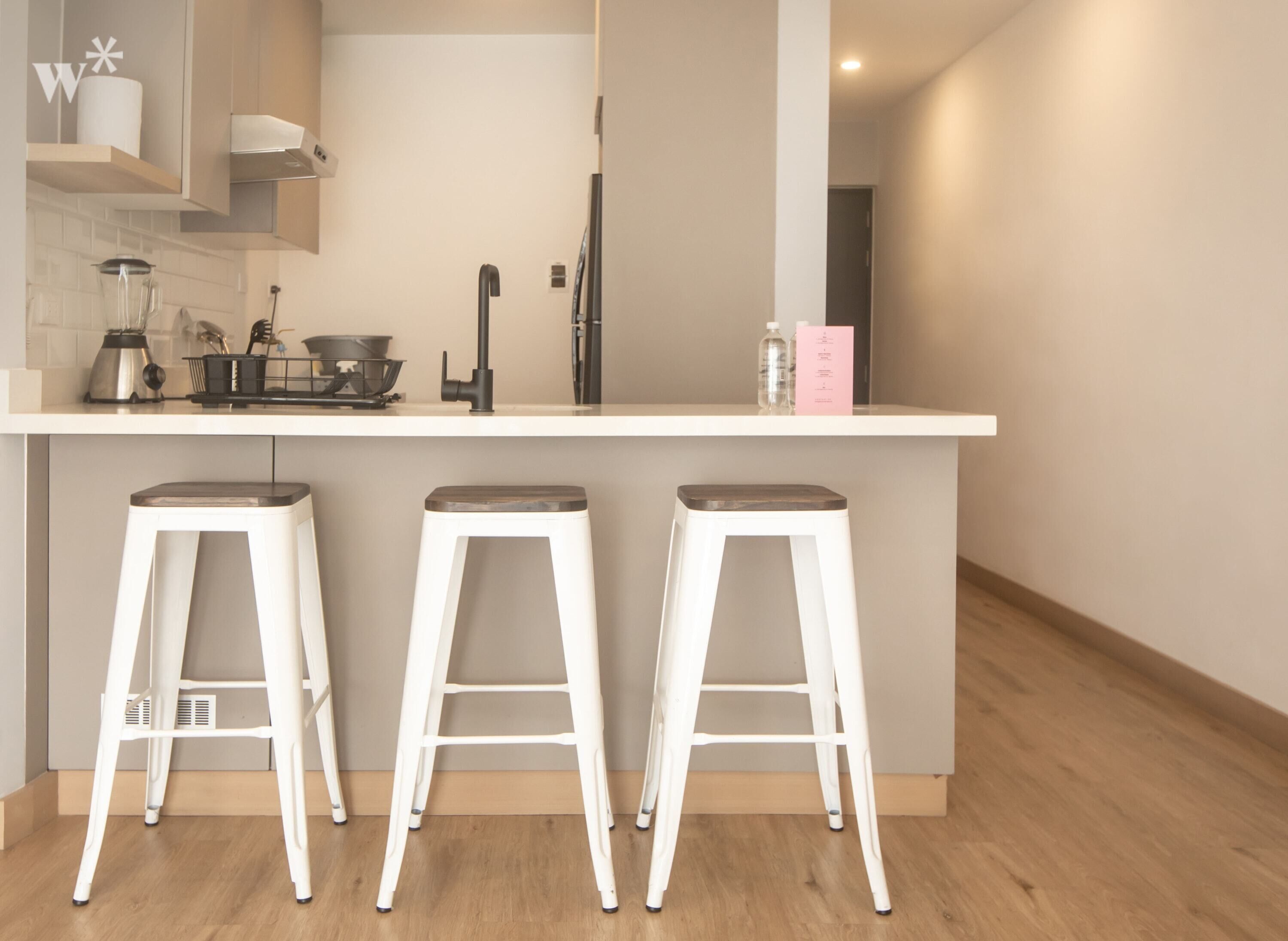Una cocina moderna con encimera blanca, tres taburetes de bar blancos y un hervidor de acero inoxidable.