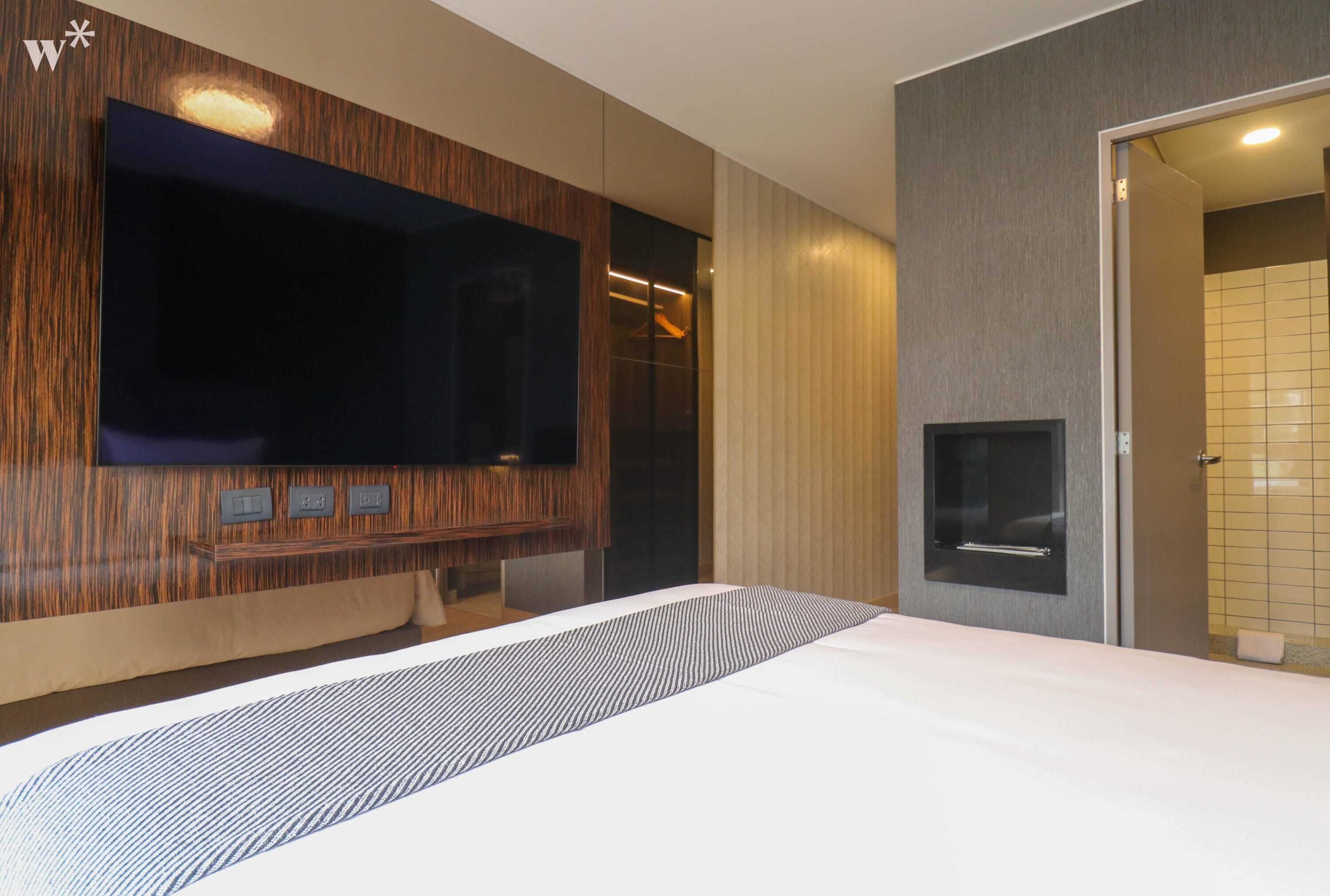 A modern hotel room with a large bed, a flat-screen TV mounted on a wooden panel, a built-in fireplace, and a shower area with tiled walls.