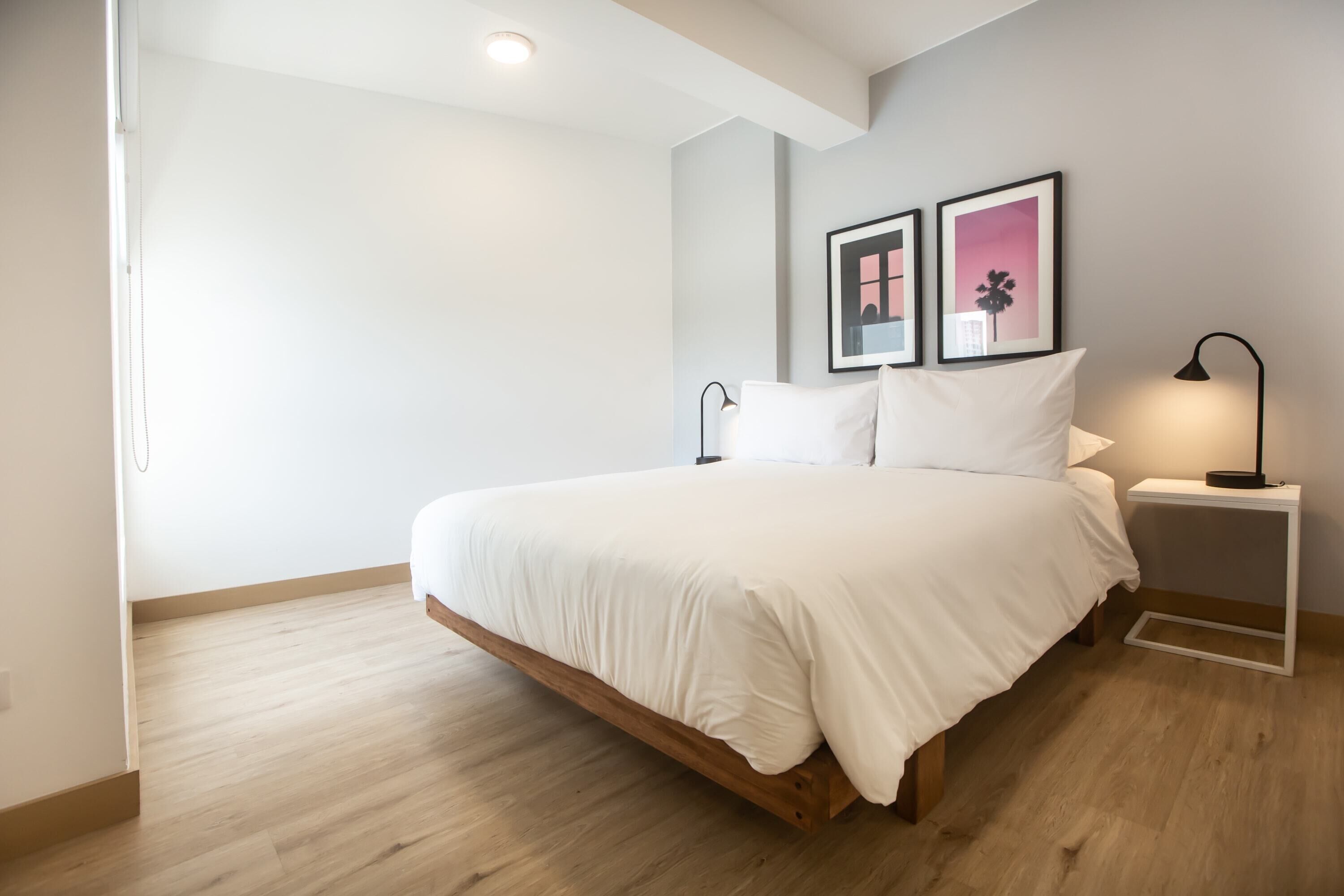 A bedroom with a wooden bed frame, white bedding, a bedside table with a lamp, and two framed pictures on the wall.