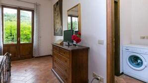 Una habitación con un aparador de madera, un monitor verde, un florero rojo, una lavadora y una ventana con vistas a follaje.