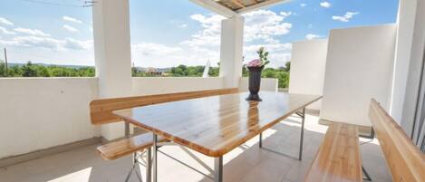 A covered outdoor patio with a wooden table and benches, large windows, and a view of greenery.