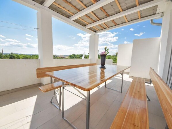 A covered outdoor patio with a wooden table and benches, large windows, and a view of greenery.