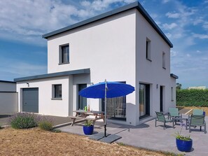 A modern two-story house with a patio area featuring a table, chairs, and a blue umbrella.