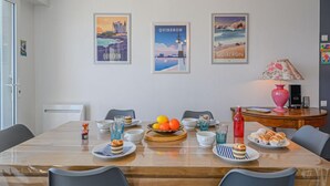 Ein Essbereich mit einem Holztisch, der für eine Mahlzeit gedeckt ist, umgeben von Stühlen. An der Wand hängen Bilder in Rahmen, auf einem Beistelltisch steht eine Lampe und im Hintergrund ist ein Kühlschrank zu sehen.