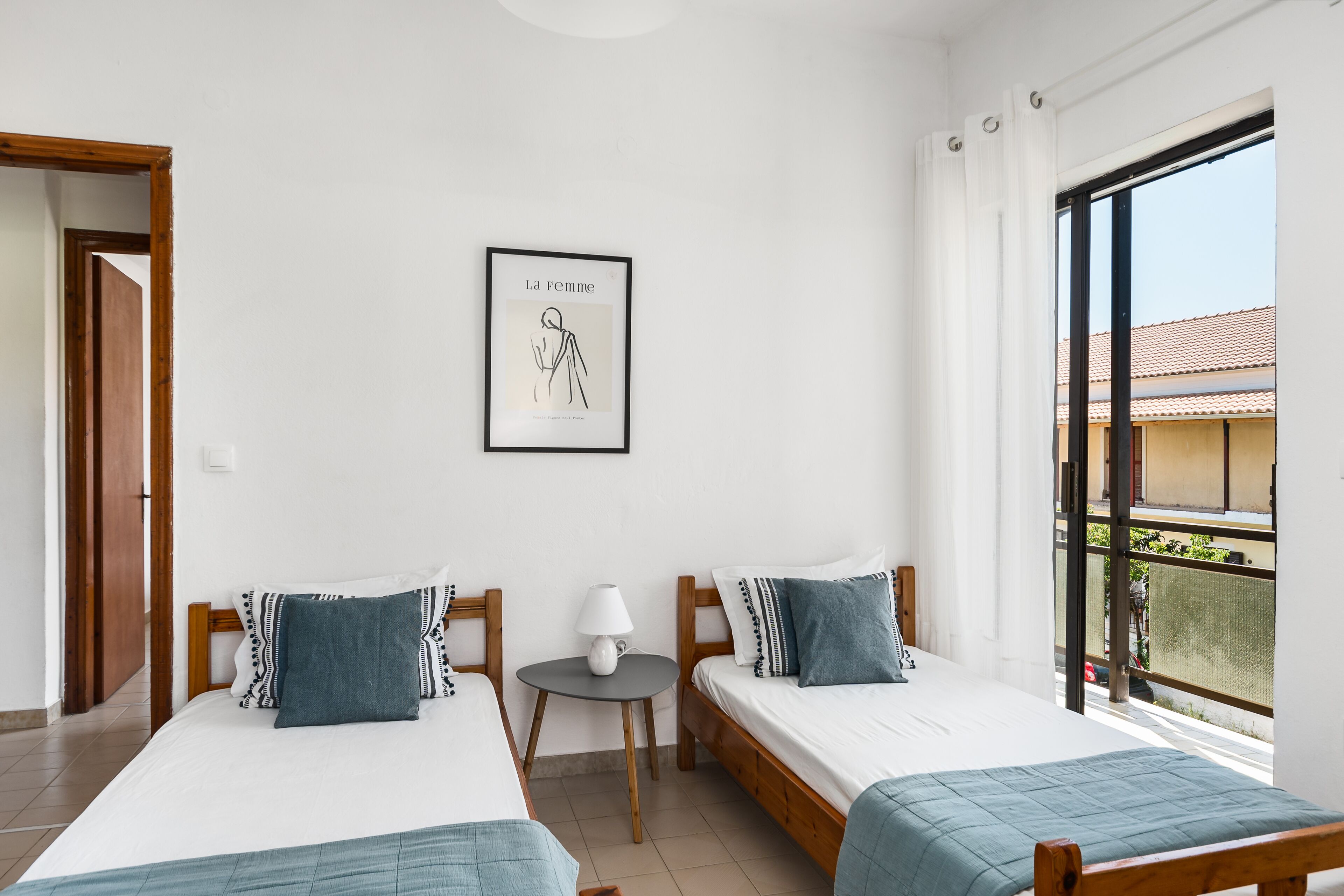 A hotel room with two beds, a small table with a lamp, a framed picture on the wall, and a large window with a view of buildings.