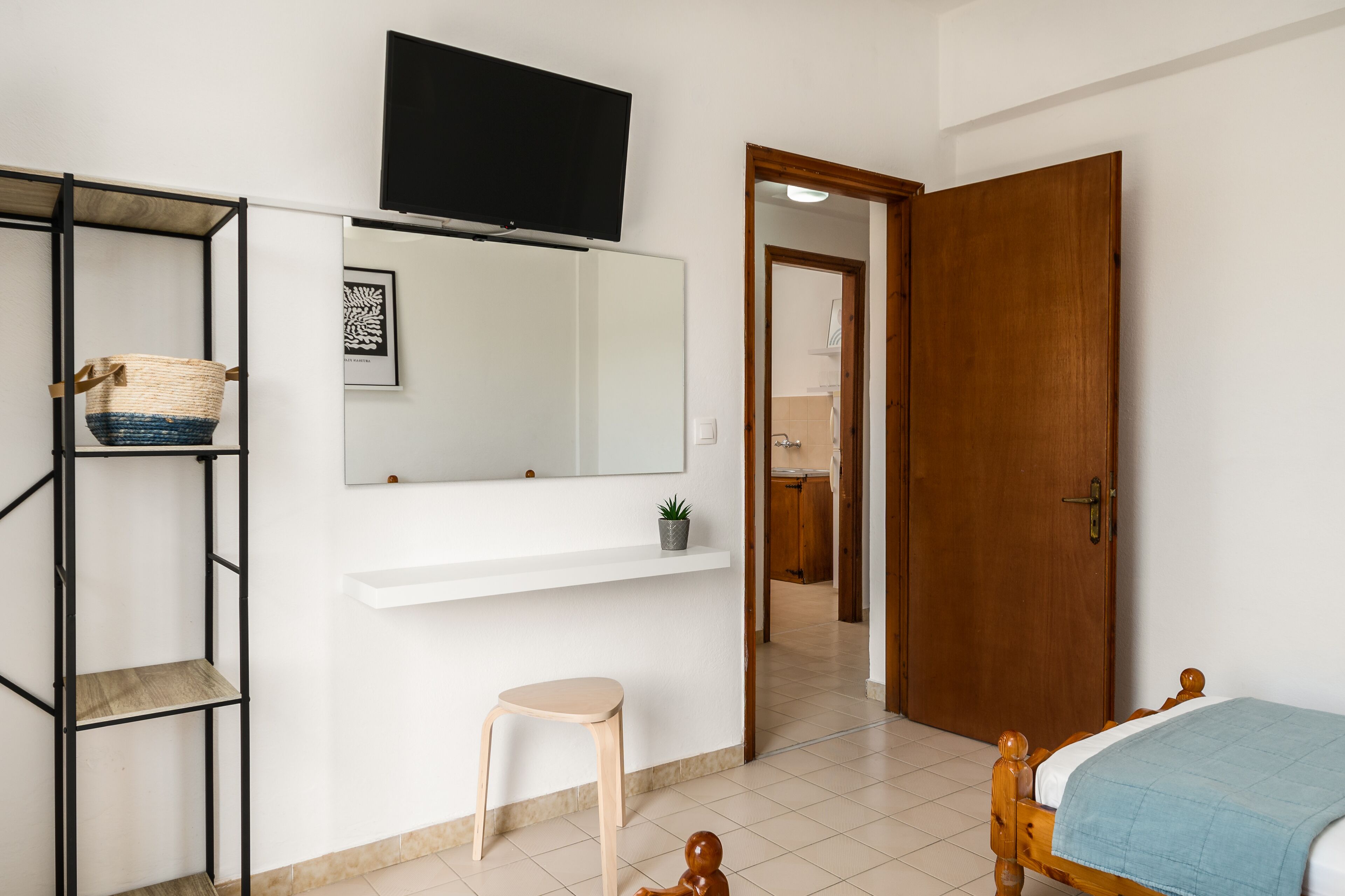 Un dormitorio con cama, puerta de madera, estante blanco, taburete pequeño, televisor y estante fijado a la pared con una cesta.