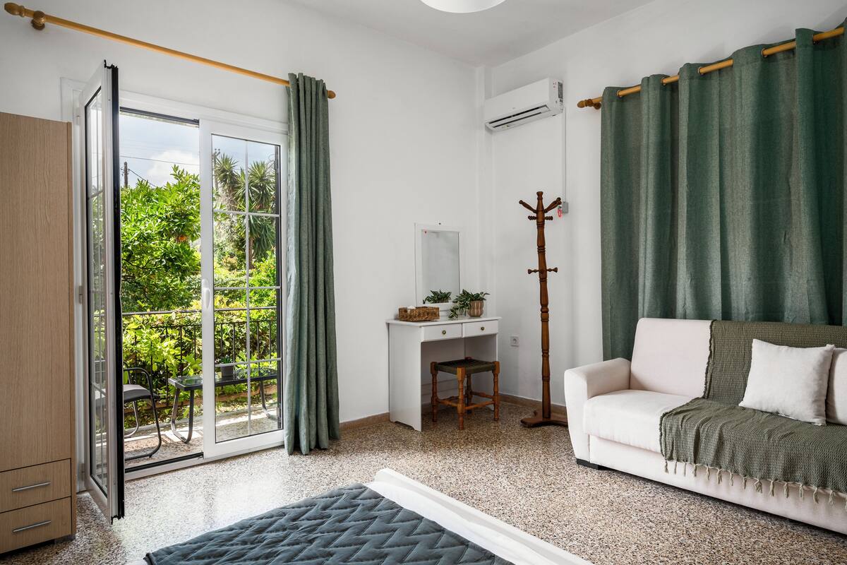 Quarto com porta de vidro deslizante para uma varanda, uma escrivaninha branca com cadeira, um sofá branco com manta verde e um cabideiro.