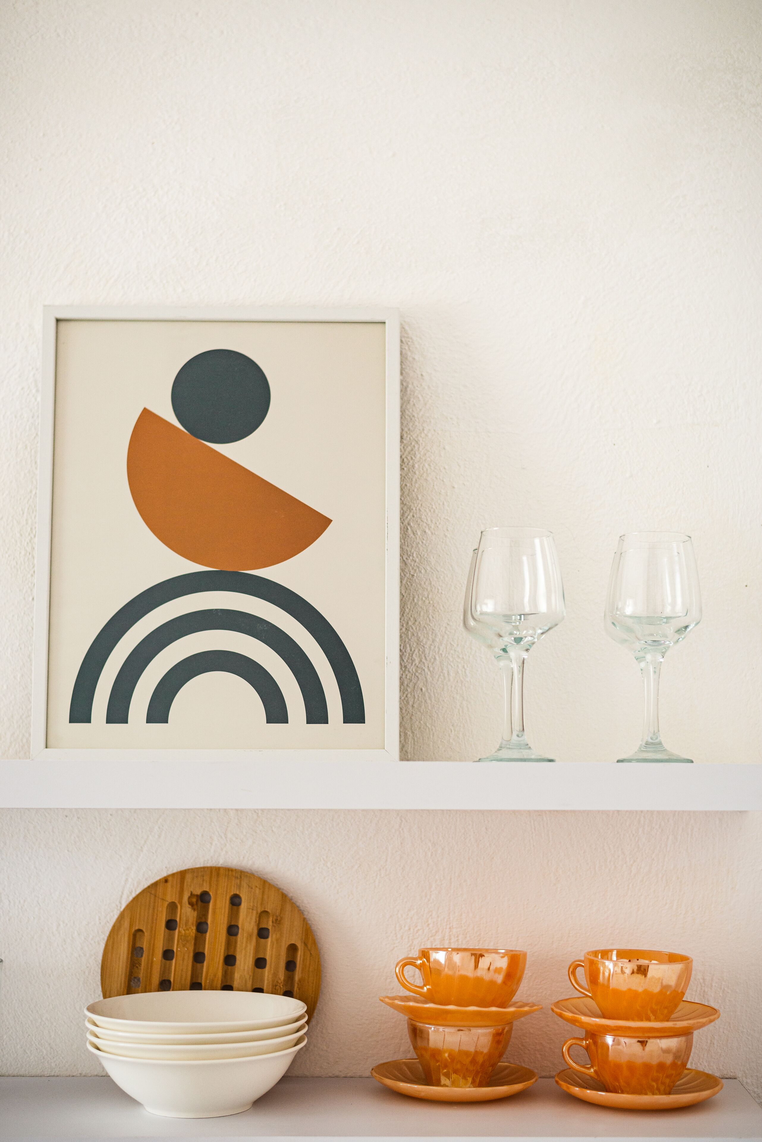 Een lijst met abstract kunstwerk, een set oranje glazen en gestapelde witte kommen op een plank.