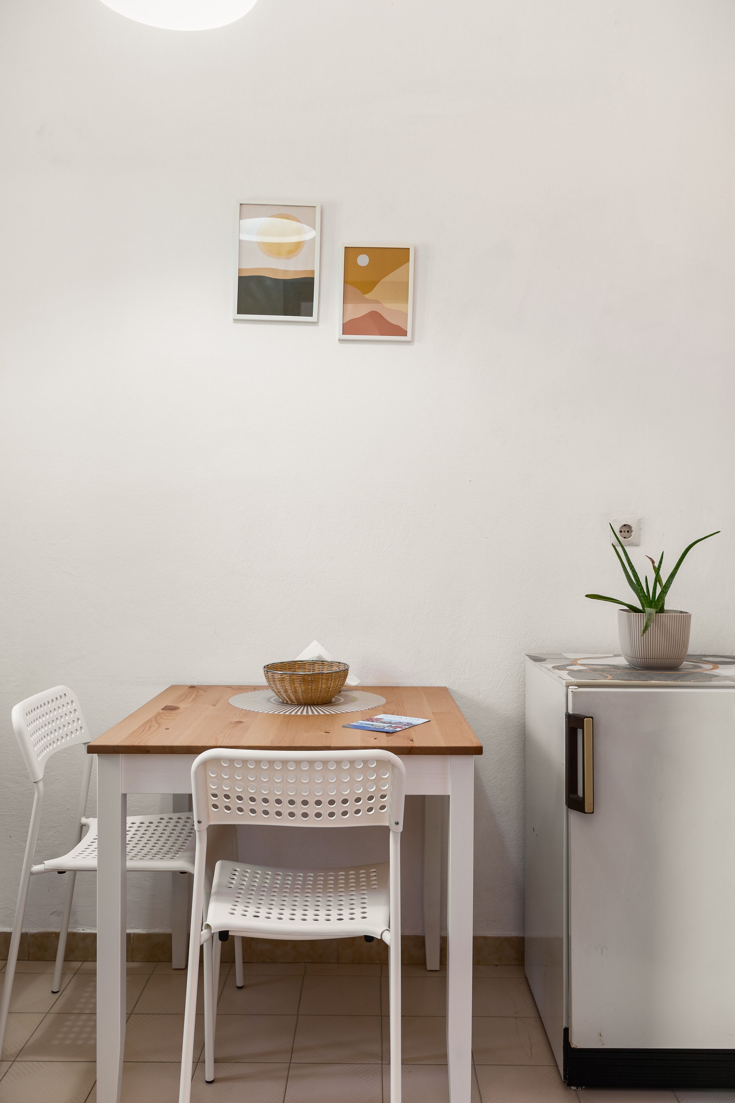 A small dining area with a wooden table, two white chairs, a wicker basket, and a potted plant on a white refrigerator.