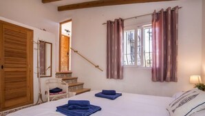Quarto com cama grande, vigas de madeira no teto, janela com cortinas, escada e porta de madeira.