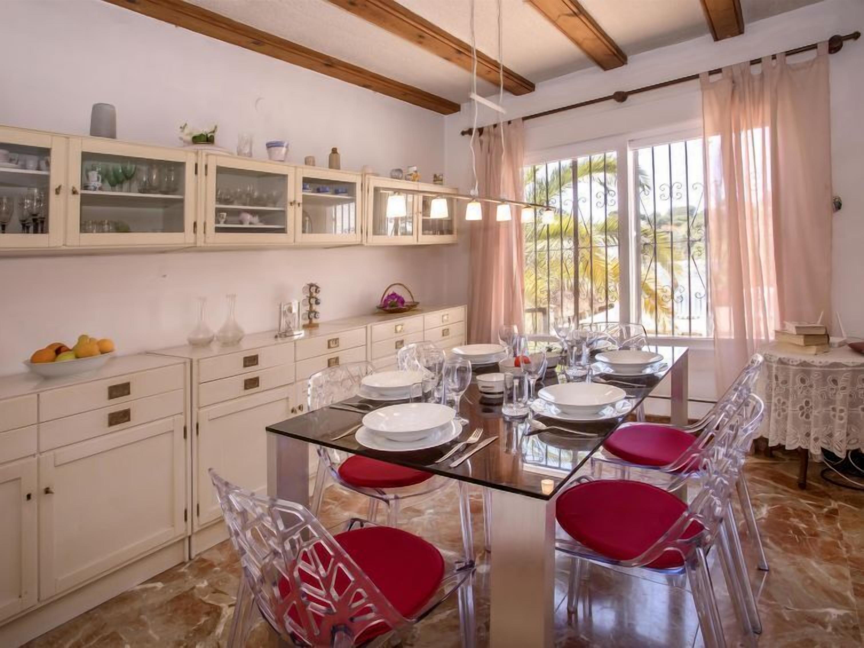A dining area with a glass table set for six, surrounded by clear chairs with red cushions. There are white cabinets with glass doors above the table, and a window with curtains to the right.