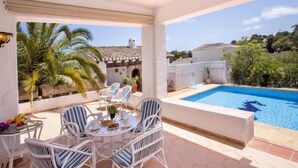 Uma área à beira da piscina com mesa e cadeiras, piscina e vista para um prédio e palmeiras.