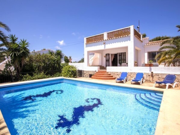 A swimming pool with blue water, surrounded by a white building with a balcony and blue lounge chairs. - Casa Moser by Interhome (Jávea)