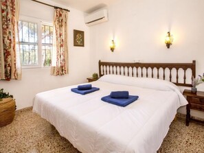 A neatly made bed with white bedding and two blue pillows, a wooden headboard, a bedside table with a lamp, and a window with floral curtains. - Casa Moser by Interhome (Jávea)