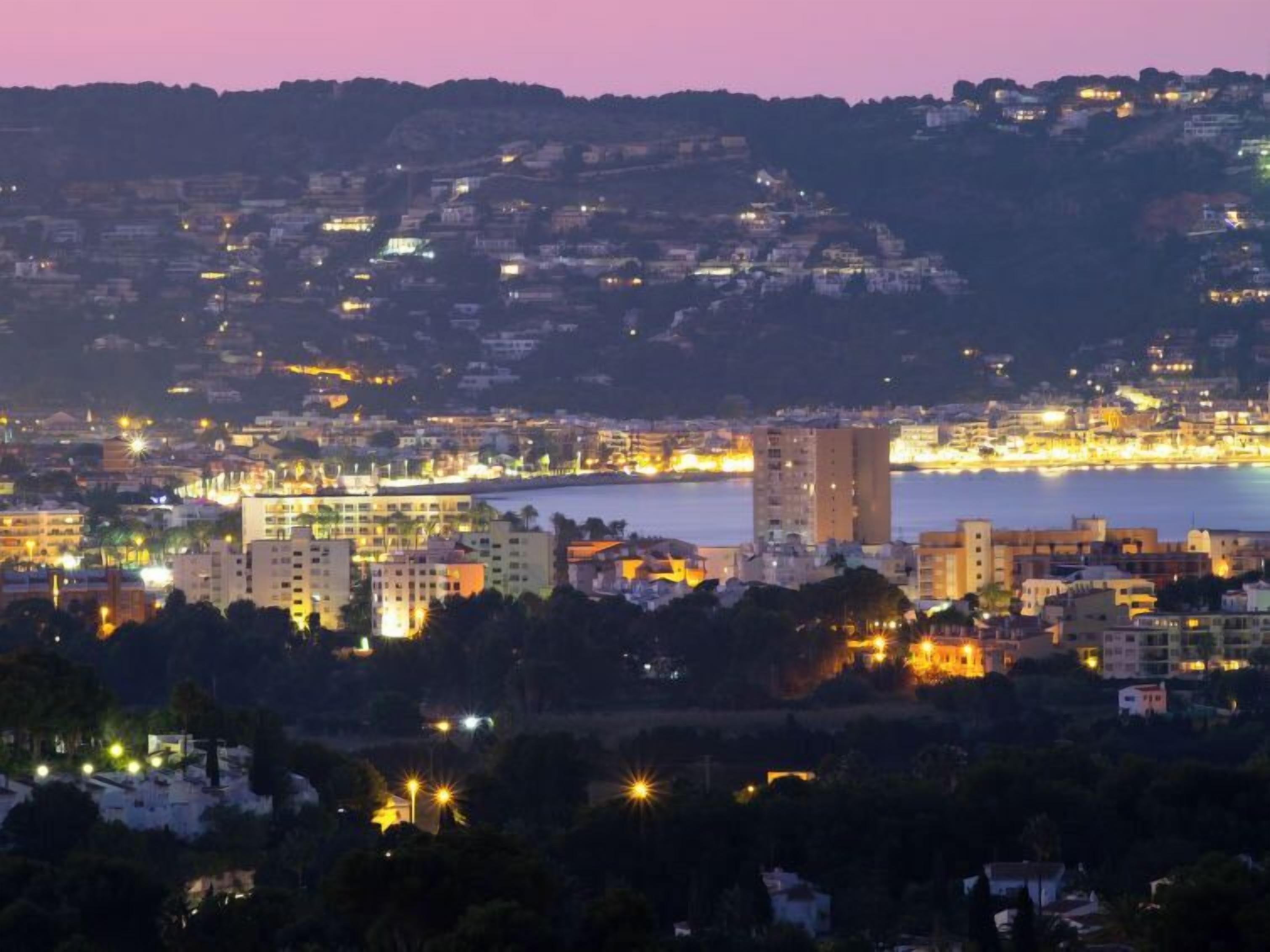 A coastal cityscape at dusk with buildings along the waterfront and a hilly backdrop.